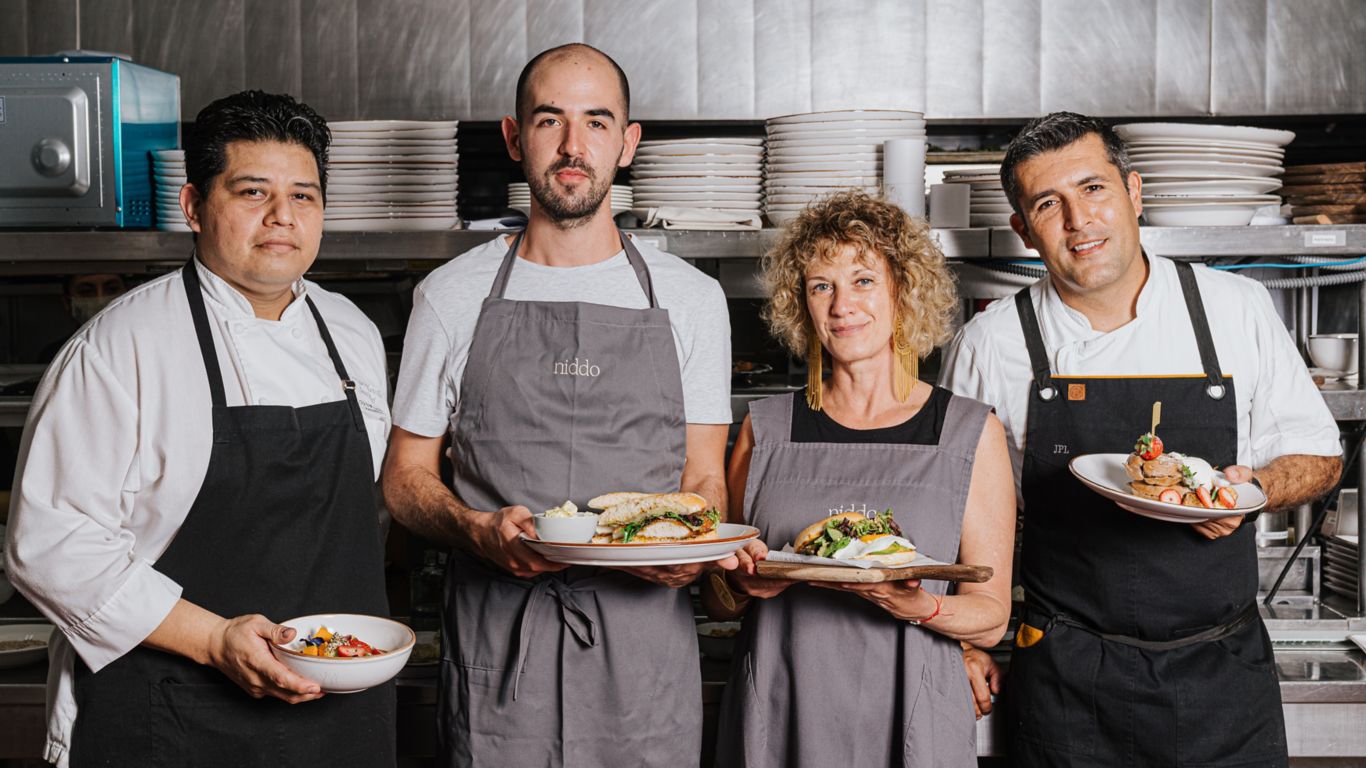 Cuatro chefs, tres hombres y una mujer en delantales de cocina de pie frente a un lugar de trabajo de cocina sosteniendo platos de desayuno. El equipo de Niddo lleva delantales grises. El equipo de Rosewood Mayakoba lleva delantales negros.